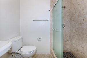 A bathroom featuring a toilet on the left and a glass shower enclosure on the right. The walls are light-colored, and the floor is tiled with a neutral pattern. A towel rack is mounted on the wall above the toilet, and the shower has a textured floor drain.
