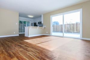 A spacious, well-lit room with a modern design featuring light-colored walls and hardwood flooring. Large sliding glass doors provide access to an outdoor patio area, and a kitchen with a breakfast bar is visible in the background. The space is open and airy, ideal for a living or dining area.