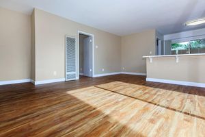 An empty room with light brown walls and a wooden floor. A doorway leads to another room, and there is a short wall or counter on the right. Natural light enters from a window, illuminating the space, creating a warm and inviting atmosphere.