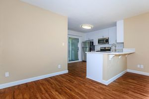 Modern kitchen and living area featuring light wood flooring, white cabinetry, stainless steel appliances, and a breakfast bar. A sliding glass door leads to an outdoor space, surrounded by beige walls and ample natural light.