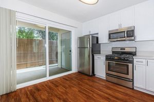 A modern kitchen featuring white cabinets, stainless steel appliances including a refrigerator and an oven, and a wooden floor. A large sliding glass door opens to a patio or balcony area with a wooden fence visible outside. Natural light fills the space, enhancing its contemporary design.