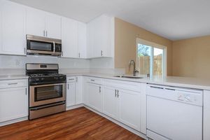 Modern kitchen featuring white cabinets, stainless steel appliances including an oven and microwave, a sink, and a dishwasher. The kitchen has a light, neutral wall color and a large window with outdoor access, providing ample natural light. The flooring is a warm wood tone.