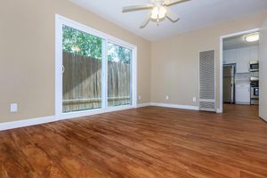A spacious room with light-colored walls and wooden flooring. Large sliding glass doors lead to an outdoor area, allowing natural light to fill the space. A ceiling fan is visible, and there is a doorway that connects to a kitchen area in the background.