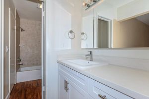 A modern bathroom featuring a white vanity with a sink, wall-mounted mirrors, and towel rings. To the left, there's a bathtub with a showerhead. The walls are light-colored, and the flooring is a warm-toned wood. The space is well-lit, creating a clean and inviting atmosphere.