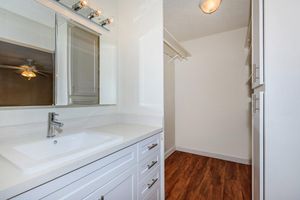 A modern bathroom featuring a white vanity with a sink, a large mirror, and stylish lighting fixtures above. The flooring is a rich wood tone, and a closet space is visible in the background, indicating an organized and well-lit area.