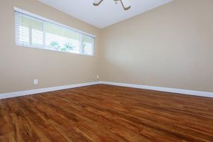 A spacious, empty room with light brown walls and a wooden floor. There's a window with white blinds, allowing natural light to enter, and a ceiling fan above. The room appears clean and ready for furniture arrangements, creating a fresh and inviting atmosphere.