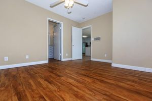 A spacious room with light brown walls and a wood-like floor. A ceiling fan hangs from the ceiling. There are two doorways leading to other areas of the home, one visible on the left and another in the back. The interior is well-lit and lacks furniture, creating an open and airy feel.