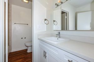 A modern bathroom featuring a white countertop sink, framed mirror, and silver towel rings. Behind the sink is a doorway leading to a separate toilet area. The walls are light-colored, and the floor has a dark wood appearance. The shower area is visible, with a clean and minimalistic design.