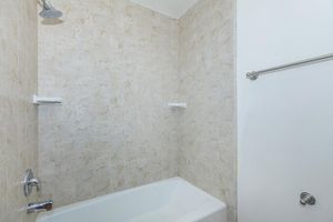 A clean, minimalist bathroom featuring a bathtub with a tiled wall. The light-colored tiles reflect a neutral palette, and there are two small shelves on the wall. A chrome showerhead is mounted above the tub, and a white shower curtain rod is installed, with the curtain not visible.