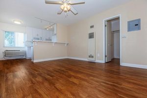 A well-lit, empty living space features light-colored walls and hardwood flooring. A ceiling fan hangs above, and there is a doorway leading to another room. The kitchen area is partially visible, showcasing a counter and white cabinetry. An air conditioning unit is mounted on the wall, providing modern comfort.