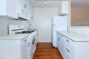 Modern kitchen with white cabinetry, a gas stove, and a microwave. Features a double basin sink and a large white refrigerator. The countertops are light-colored with a wooden floor, creating a clean and inviting space. Well-lit with overhead lighting, the area is organized and contemporary in design.