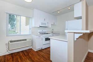 Modern kitchen featuring white cabinetry, a gas stove, and microwave. There’s a refrigerator partially visible, and an air conditioning unit along the wall. Natural light comes through a window with blinds, illuminating the wood-style flooring and minimalist design.