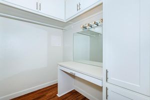 A well-lit dressing area featuring a large mirror, a white countertop, and storage cabinets above. The space has light-colored walls and wood flooring, providing a clean and modern look. Ideal for a vanity or makeup station.
