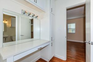 A bright room features a white vanity with a mirror and overhead lights, flanked by storage cabinets. A doorway leads to another room with a soft beige wall and a window, illuminating the wooden floor. The design is clean and modern, emphasizing simplicity and functionality.