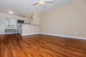 A spacious, empty living area featuring light brown wooden flooring, a ceiling fan, and a partially open kitchen with a bar. The kitchen includes modern appliances, and natural light comes in through a nearby window. The walls are painted in a soft beige color, creating a warm atmosphere.