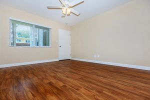 A spacious, empty room featuring light-colored walls and a wooden floor. There's a window with blinds allowing natural light to enter, and a ceiling fan above. A closed door is visible on one wall, emphasizing the room's simplicity and potential for personalization.
