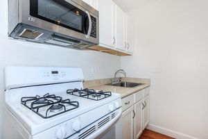 Compact kitchen featuring a white gas stove, stainless steel microwave mounted above, and a sink with a countertop. There are white cabinets above the countertop, and the flooring is a wood-like finish. The walls are painted in a light color, creating a bright and clean appearance.