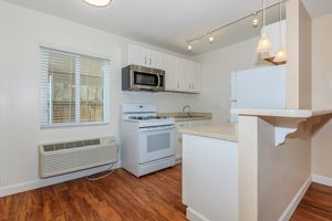 A modern kitchen featuring white cabinetry, stainless steel appliances including a microwave and gas stove, and a white refrigerator. The space has wooden flooring, a window with blinds, and pendant lighting over the counter, creating a bright and inviting atmosphere.