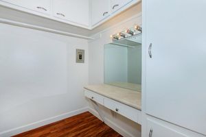 A well-organized, empty dressing area featuring a mirror, light fixtures above, and a countertop. The space has white cabinetry and a warm wooden floor, creating a bright and clean aesthetic. The walls and ceiling are painted in a light color, enhancing the openness of the room.