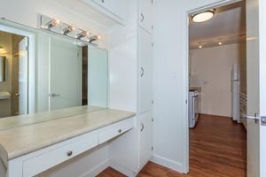 A brightly lit bathroom featuring a large mirror above a countertop with drawers. The walls are painted a light color, and there is wood flooring. A doorway leads to a kitchen area in the background, which includes white appliances. The space is tidy and modern, with ample natural light.