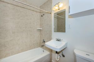 A small bathroom featuring a light-colored tiled wall, a bathtub with a showerhead, a wall-mounted sink, and a mirror above the sink. A light fixture is fixed to the wall near the ceiling, and a compact toilet is situated beside the sink. The overall design is simple and functional.