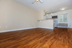 A spacious, unfurnished living area with light-colored walls and modern wooden flooring. A small kitchen area is visible in the background, featuring white cabinetry and appliances. Soft lighting from overhead fixtures enhances the bright, open feel of the room.