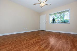 A spacious, empty room with light-colored walls and a wooden floor. A ceiling fan is visible, and sunlight streams through a window with blinds. The door is white, and there are no furnishings, creating a minimalist and inviting atmosphere.