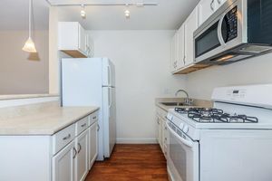 A bright kitchen featuring white cabinetry, a gas stove, a microwave above it, and a refrigerator. Countertops are light-colored, and the floor is wood laminate. There are overhead light fixtures and the space is well-organized, creating an inviting cooking environment.