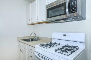 A small kitchen featuring white cabinetry, a stainless steel microwave above the stove, and a white gas range. There is a stainless steel sink and countertop. The walls are painted in a light color, giving the space a clean and modern feel.
