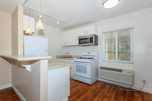 A bright, modern kitchen featuring white cabinetry, a gas stove, microwave, and refrigerator. The space has a light-colored countertop and wood-like flooring. There are two pendant lights above the kitchen counter and a window providing natural light. An air conditioning unit is visible on the wall.