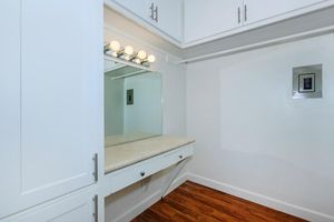 A well-lit vanity area with a mirror and a countertop, featuring five lightbulbs above. The walls are white, with a built-in cabinetry design. The floor is wooden, contributing to a warm ambiance. A small wall-mounted panel is visible nearby.