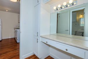 A well-lit bathroom area featuring a vanity with a large mirror and light fixtures above. The background shows part of a kitchen with white cabinetry and a stove. The floor is made of wood, and the walls are painted in a light color, creating a spacious and bright atmosphere.