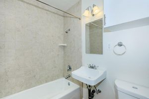 A small bathroom featuring a bathtub with a shower, a wall-mounted sink, a mirror, and a toilet. The walls are tiled in a light color, and there is a small shelf beside the showerhead. The lighting is provided by two wall-mounted fixtures above the mirror.