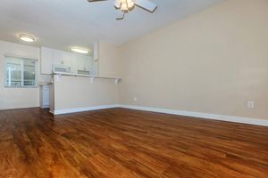 A spacious room with light-colored walls and a wooden floor. The area features a ceiling fan and a bar counter leading to a kitchen. Natural light comes through a window, creating a warm atmosphere. The room is empty, providing an open and inviting feel for future furnishings.