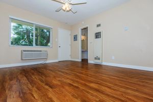 A well-lit, empty room featuring a ceiling fan, a window with blinds, and light beige walls. The floor is made of polished wood, and there's an air conditioning unit on one wall. A doorway leads to a bathroom visible in the background, creating a simple and inviting space.