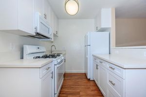 Modern kitchen featuring white cabinetry, a gas stove with an oven, a microwave, and a refrigerator. The countertops are light-colored, and the flooring is wooden. The area is well-lit with a circular ceiling light, creating a bright and inviting ambiance.