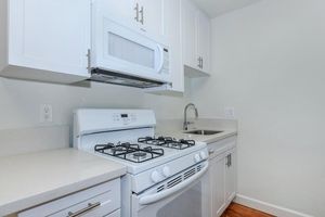 A modern kitchen featuring white cabinetry, a gas stove with four burners, a microwave above it, and a sink. The countertop is smooth and light-colored, while the floor is made of hardwood. The overall design is clean and minimalist, with a bright and airy feel.