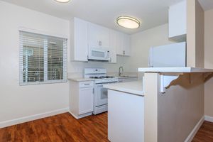 A modern, clean kitchen with white cabinets, a gas stove, a microwave, and a sink. There's a window with blinds providing natural light, and the floor is a warm wood-tone laminate. The layout features an open counter space, creating a welcoming and functional environment.