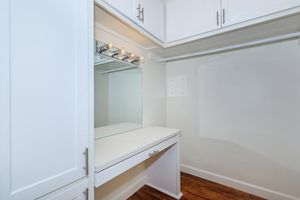 A well-lit vanity area in a closet featuring a white countertop and a large mirror with four light fixtures above. The surrounding cabinetry is white with modern handles, and the floor is a warm wood laminate. The space appears organized and functional with ample room for storing personal items.