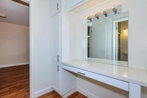 A bright and modern vanity area featuring a large mirror mounted above a white countertop with vanity lights. The surrounding cabinetry is sleek and white. The room has light-colored walls and warm wooden flooring, with an adjacent doorway leading to another space, creating a spacious and inviting atmosphere.