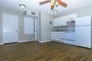 A small kitchen area featuring white cabinets, a white stove, and a refrigerator. The floor is wooden, and the walls are painted in a light gray color. A ceiling fan is visible, and there is a door leading to another room. Sunlight enters through the doorway, creating a bright atmosphere.