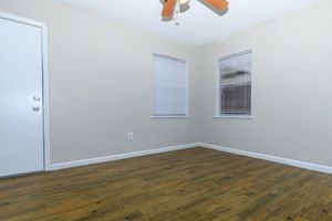 Empty room with light gray walls and a wooden floor. There is a white door on the left and two windows with blinds on the right. A ceiling fan with wooden blades is visible above, and the space appears well-lit and clean.