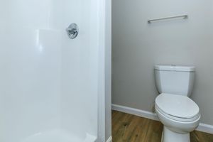 A clean and modern bathroom featuring a white toilet next to a shower stall with a plain, white interior. The floor is wood-like, and the walls are light-colored, giving a spacious and bright appearance. A towel bar is mounted on the wall above the toilet.