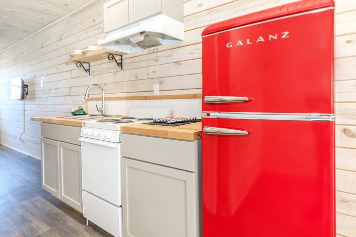 A bright and modern kitchen featuring a striking red refrigerator, light wood paneling, and sleek gray cabinetry. The kitchen includes a white stove, a sink, and open shelving above the counter, adding a contemporary touch to the cozy space.