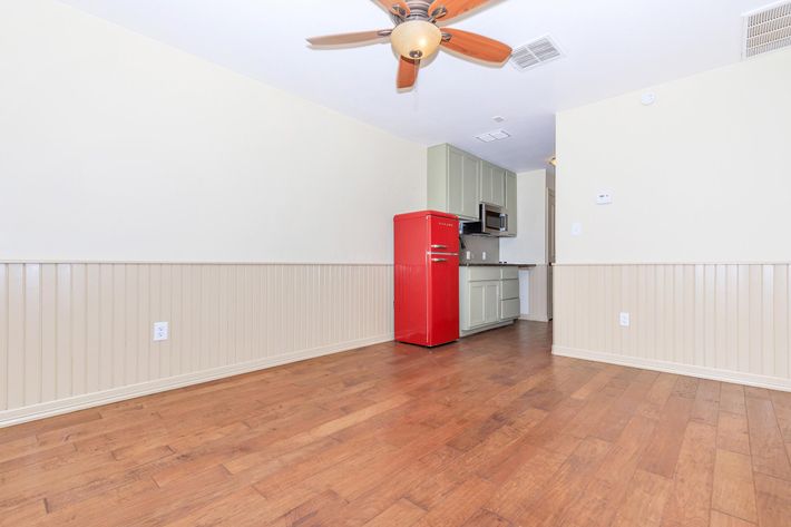 An empty room with hardwood flooring and a ceiling fan. On the left, a bright red refrigerator stands against a light green kitchen area. The room features light-colored walls with wainscoting. A kitchen countertop and cabinet are visible in the background, creating a cozy, modern ambiance.