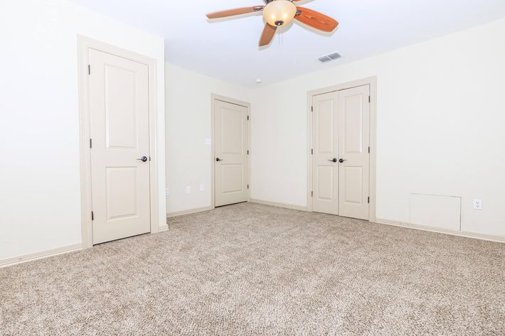 An empty room with beige walls and a ceiling fan. There are two closed doors on the left and one on the right, all matching in design. The floor is covered in light brown carpet, and the room appears well-lit with natural light. No furniture or decorations are present.
