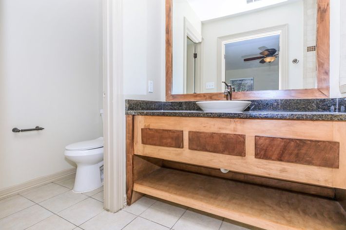 A clean, modern bathroom featuring a wooden vanity with a sink, a large mirror, and a ceiling fan. The floor is tiled, and there is a toilet visible in the background. The space has neutral-colored walls and a minimalistic design.
