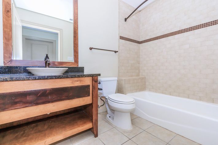 A clean and modern bathroom featuring a white bathtub, a toilet, and a wooden vanity with a dark countertop and sink. The walls are tiled in light beige, with a decorative border tile. Natural light enters from an unseen door, adding brightness to the space.