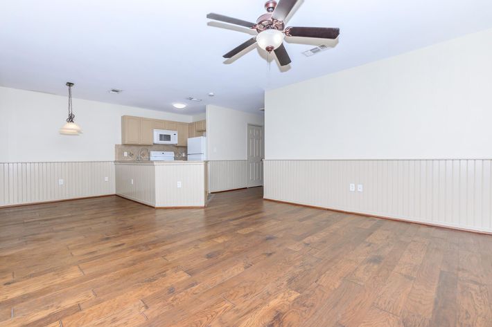 A spacious living area with hardwood floors, a ceiling fan, and light-colored walls. The room features a kitchen area with a white microwave and refrigerator visible in the background, alongside a pendant light hanging from the ceiling. The space is bright and inviting, suitable for various furnishings.