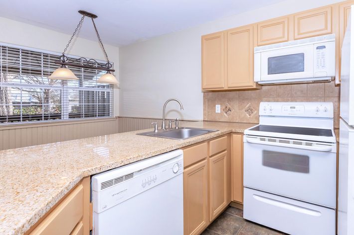 Modern kitchen featuring light-colored cabinets, a white stove, and built-in microwave. Includes a stainless steel sink with a brushed metal faucet, a dishwasher, and a spacious granite countertop. The space is illuminated by pendant lighting and has a window providing natural light. Decorated in a clean, contemporary style.